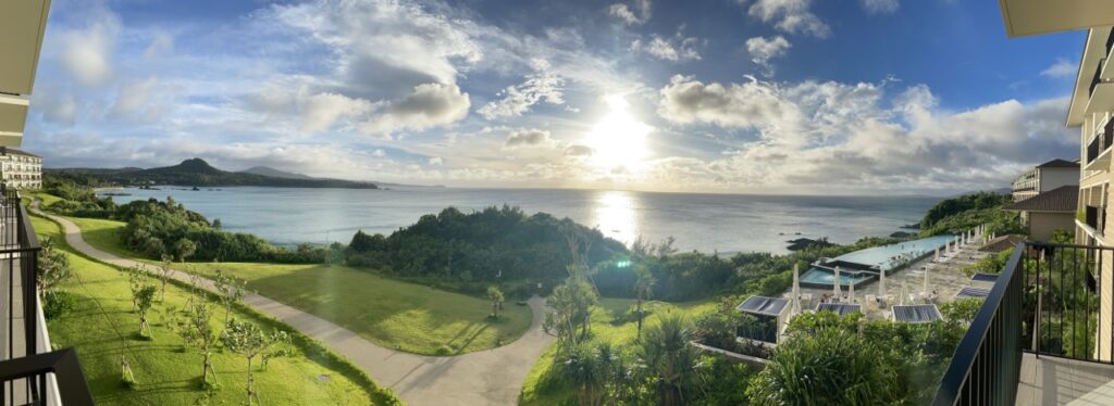 ハレクラニ沖縄の客室バルコニーから望む東シナ海パノラマビュー。インフィニティプールと緑の庭園、朝日に輝く海が広がる