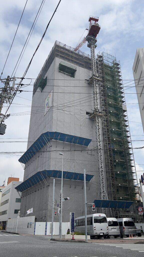 2026年4月時点のKOKO HOTEL 那覇前島（仮称）の建設現場。外壁の大部分が養生シートで覆われ、屋上には赤いタワークレーンが設置されている様子。