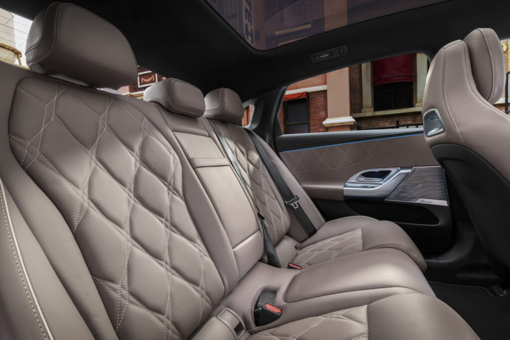 新型電動Cクラス（2026年）リアシート。ダイヤモンドキルティングのベージュ調ヴィーガンレザーと白ステッチ、ドアパネルのBurmesterスピーカーグリルおよびブルーアンビエントライト